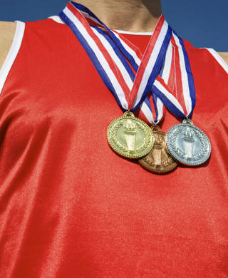 Athlete in red athletic shirt stands with gold, silver, and bronze medals in front of bright blue sky